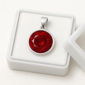 May include: A round, red gemstone pendant with a silver-colored bezel and bail. The pendant is set in a white jewelry box. The gemstone has multiple facets, reflecting light. The jewelry is displayed on a white background.