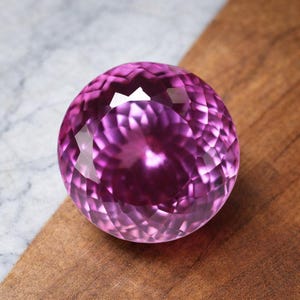 May include: A faceted, spherical gemstone in shades of purple and pink. The gem is highly polished, reflecting light and creating a brilliant display. It sits on a wooden surface, with a marble background.