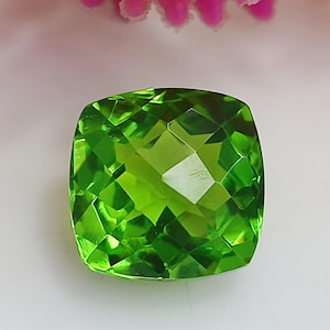 Puede incluir: Una gema verde vibrante con un corte cojín y un diseño facetado. La gema es clara y tiene un alto nivel de brillo.