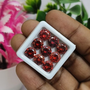 Pode incluir: Uma caixa branca com uma tampa transparente contendo nove pedras preciosas vermelhas. As pedras preciosas são redondas e têm um corte facetado.