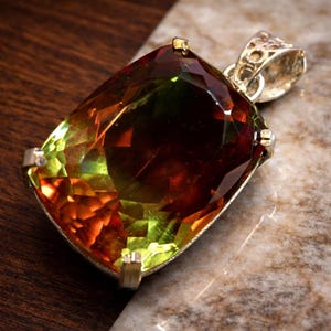 May include: A large, rectangular pendant with a faceted gemstone. The gem displays a blend of colors, including shades of green, orange, and brown. The pendant is set in a silver-colored metal with a decorative bail, suitable for a necklace.