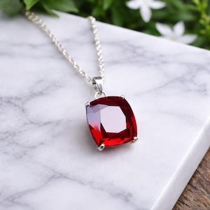 May include: A silver necklace with a large, square-cut, ruby-red gemstone pendant. The pendant is set in a silver bezel and hangs from a delicate silver chain. The gemstone has multiple facets, reflecting light. The necklace is displayed on a white marble surface.