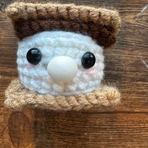 Puede incluir: Un juguete de s'more hecho a mano con ganchillo con una cara de malvavisco blanco, ojos de botón negros y una nariz blanca. El s'more tiene una parte superior e inferior de galleta Graham marrón y una base de galleta Graham beige.