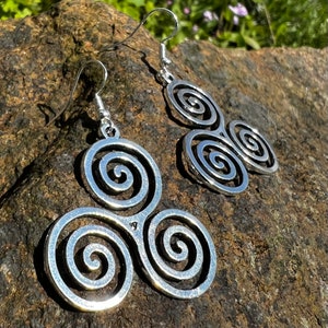May include: Silver-toned spiral earrings. Each earring features a triple spiral design, with a hook for easy wear. The earrings are displayed on a textured, dark brown surface, with natural light enhancing their metallic sheen. The design is Celtic-inspired.