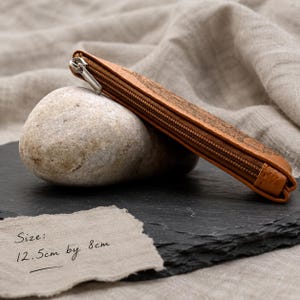 May include: A tan leather pencil case with a silver metal zip closure and an embossed floral pattern. The case measures 12.5 cm by 8 cm as indicated by the text. The item is positioned at an angle, resting against a smooth, light-coloured stone.