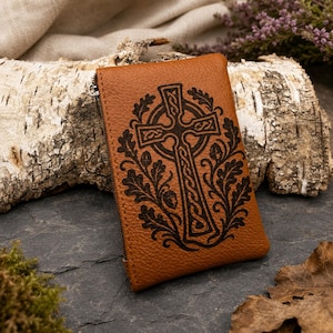 May include: A tan, textured leather pouch featuring a dark, laser-engraved Celtic cross design surrounded by intricate oak leaf and acorn patterns. The rectangular accessory includes a side zip closure and a visible metal zip pull.