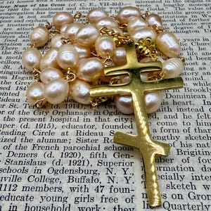 Puede incluir: Un collar con colgante de cruz dorada y una cadena de perlas ovaladas blanquecinas. La cruz tiene un diseño de doble barra. El collar se muestra sobre una página de libro antiguo.