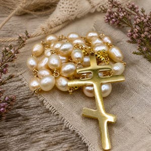 May include: A gold Orthodox cross pendant with a double bar design, strung with creamy, irregular-shaped pearls. The necklace is displayed on a neutral-toned linen cloth, with sprigs of dried flowers in the background.
