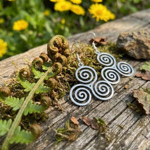 May include: Silver Celtic spiral earrings displayed on a wooden surface with moss and ferns. The earrings feature a triple spiral design, a common Celtic symbol. The background includes yellow flowers and natural elements.