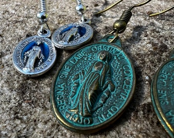 May include: Close-up of three pairs of earrings. Two pairs have a silver finish with a blue background and the words "REGINA SINE LABE ORIGINAL CONCEPTA OPN" around a religious figure. The third pair has a bronze finish with the same text and figure.
