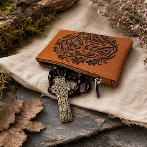 May include: A tan leather zippered pouch featuring a dark brown embossed Celtic cross and oak leaf design. A set of dark brown wooden prayer beads with a metallic bronze-toned Celtic cross pendant hangs from the open zipper of the pouch.