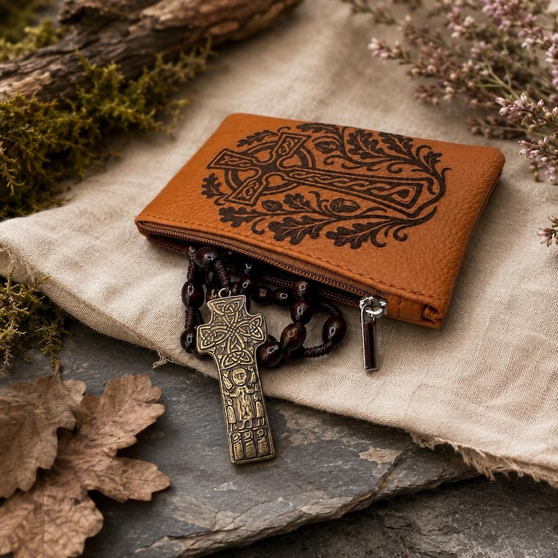 May include: A tan leather zippered pouch featuring a dark brown embossed Celtic cross and oak leaf design. A set of dark brown wooden prayer beads with a metallic bronze-toned Celtic cross pendant hangs from the open zipper of the pouch.