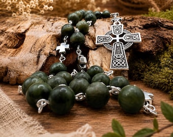May include: A rosary with dark green, round beads and silver-toned metal accents. The rosary features a Celtic cross pendant and smaller cross-shaped connectors. The beads are strung on a silver chain, and the rosary rests on a wooden surface.