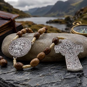 Könnte beinhalten: Ein Rosenkranz mit braunen Holzperlen auf hellem Kordelband, einer runden silberfarbenen Medaille mit der Darstellung des heiligen Christophorus und einem silberfarbenen, verzierten Kreuz mit detaillierten Reliefmustern.