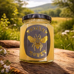Könnte beinhalten: Ein klares Glasgefäß gefüllt mit hellem Honig, versehen mit einem schwarzen sechseckigen Deckel. Das vordere Etikett ist ein dunkelgraues Oval mit einer goldenen Bienenillustration und dem Text "PAISLEY HONEY" in dekorativer Goldschrift.