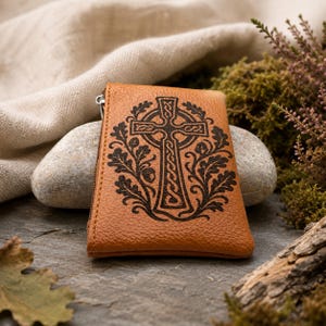 May include: A tan leather coin purse featuring a dark brown laser-engraved design of a Celtic cross surrounded by oak leaves and acorns. The rectangular pouch has a side zipper closure and a textured surface, showcasing detailed knotwork patterns.