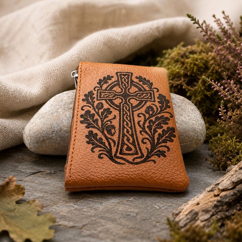 May include: A tan leather coin purse featuring a dark brown laser-engraved design of a Celtic cross surrounded by oak leaves and acorns. The rectangular pouch has a side zipper closure and a textured surface, showcasing detailed knotwork patterns.