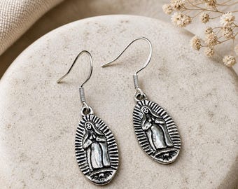 May include: A pair of silver-colored dangle earrings. Each earring features an oval pendant with a religious figure in the center, surrounded by a radiating design. The earrings have a hook closure.
