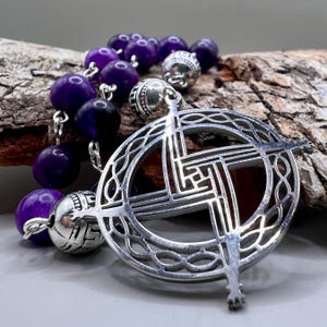 Puede incluir: Un colgante de nudo celta de plata con un collar de cuentas de amatista púrpura. El colgante tiene un diseño circular con una cruz en el centro.