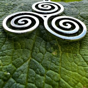 May include: A silver-colored pendant featuring a Celtic triskele design, composed of three interconnected spirals. The pendant is displayed on a textured green leaf, creating a contrast between the metal and the natural background.