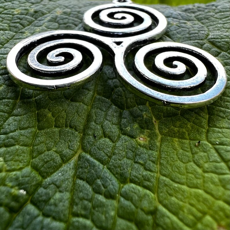 May include: A silver-colored pendant featuring a Celtic triskele design, composed of three interconnected spirals. The pendant is displayed on a textured green leaf, creating a contrast between the metal and the natural background.