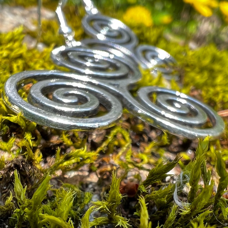 May include: A silver-colored Triskele pendant earring with three spiral arms. The earring is set against a backdrop of green moss and yellow flowers, creating a natural and earthy aesthetic.
