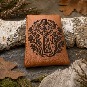May include: A tan leather pouch featuring a dark brown embossed design of a Celtic cross surrounded by oak leaves and acorns. The rectangular pouch has a visible silver-toned metal zipper pull on the left side and a textured grain surface.