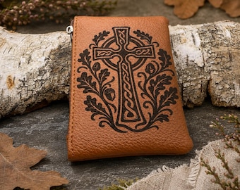 May include: A tan leather pouch featuring a dark brown embossed design of a Celtic cross surrounded by oak leaves and acorns. The rectangular pouch has a visible silver-toned metal zipper pull on the left side and a textured grain surface.