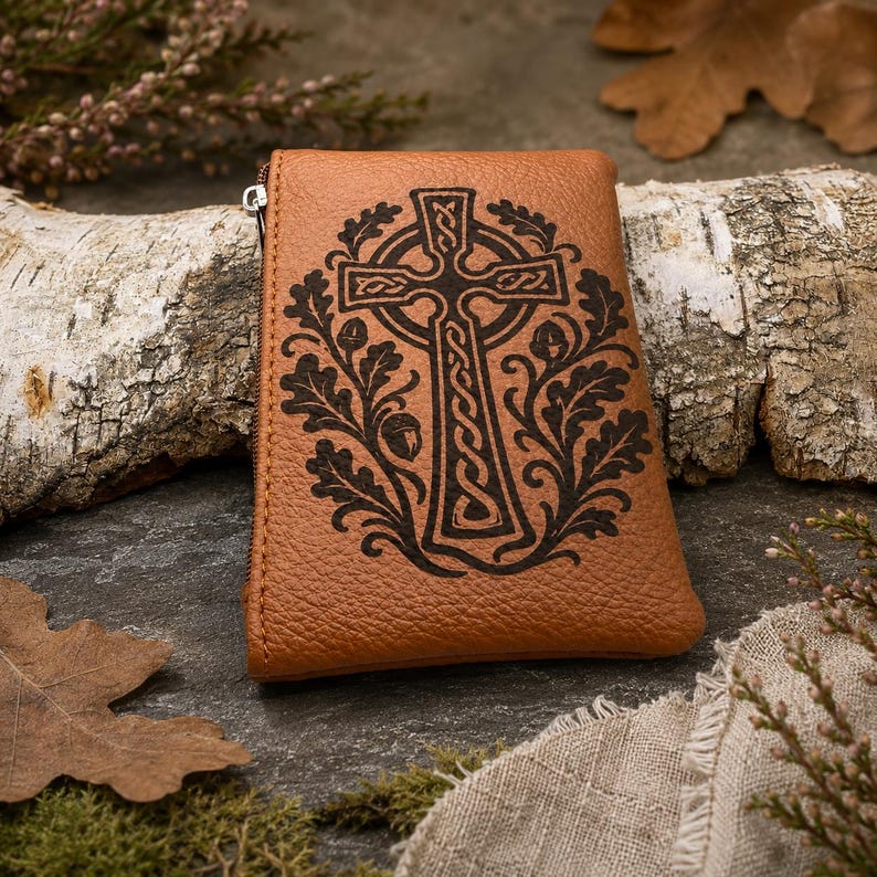 May include: A tan leather pouch featuring a dark brown embossed design of a Celtic cross surrounded by oak leaves and acorns. The rectangular pouch has a visible silver-toned metal zipper pull on the left side and a textured grain surface.