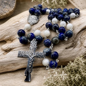 May include: A rosary with alternating blue and white beads. The beads are strung on a silver chain, leading to a detailed silver cross. The cross has a tree-like design. The rosary is displayed on a piece of driftwood.
