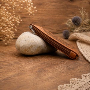 May include: A tan leather zippered pouch with a dark embossed pattern resting against a smooth, light grey oval stone. The pouch features a silver-toned metal zipper pull and a slim, rectangular profile with visible stitching along the edges.