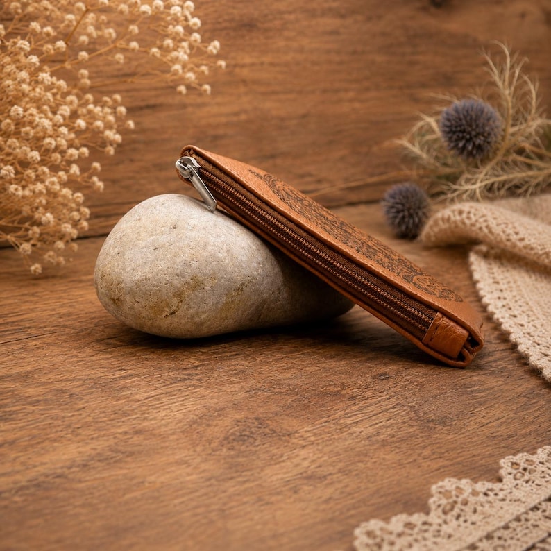 May include: A tan leather zippered pouch with a dark embossed pattern resting against a smooth, light grey oval stone. The pouch features a silver-toned metal zipper pull and a slim, rectangular profile with visible stitching along the edges.