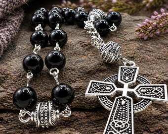 May include: A rosary featuring smooth, round black beads connected by silver-toned metal links. The piece includes decorative silver-toned spacers and a large, detailed Celtic cross pendant with intricate knotwork patterns and a textured finish.