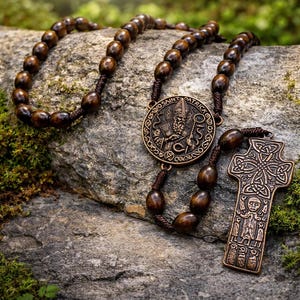 May include: A wooden rosary with dark brown beads and a bronze Saint Patrick medallion and Celtic cross pendant. The rosary is displayed on a gray rock with green moss.