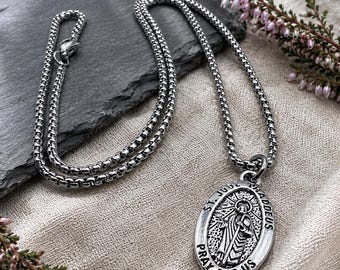 Op de afbeelding: Een zilverkleurige metalen ketting met een box chain en een ovaal hangertje. De hanger toont een reliëfafbeelding van een figuur met een staf, omringd door de tekst ST. JUDE THADDEUS PRAY FOR US. De ketting heeft een karabijnsluiting.