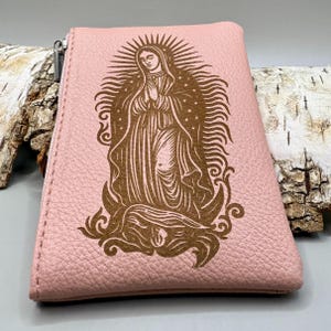 May include: A pink leather coin purse with a zipper closure. The purse features a detailed brown illustration of the Virgin Mary with a halo and praying hands. The design is centered on the front of the purse.