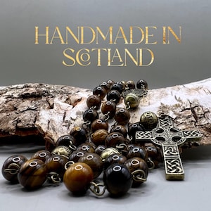 Puede incluir: Un rosario de cuentas de ojo de tigre marrón y dorado con un colgante de cruz celta. El rosario está hecho a mano en Escocia.