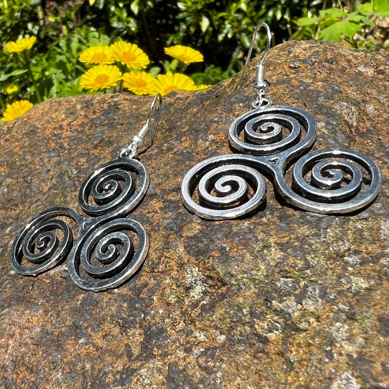 May include: Silver-toned spiral earrings. Each earring features a design of three interconnected spirals. The earrings are displayed on a textured, dark brown rock, with yellow flowers and green foliage in the background.