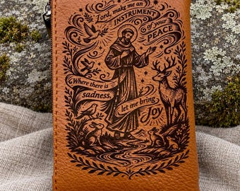 May include: A tan leather pouch featuring a dark brown laser-engraved illustration of Saint Francis of Assisi surrounded by animals and nature. The text reads: Lord, make me an INSTRUMENT of your PEACE. Where there is sadness, let me bring JOY.