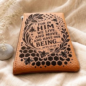 Peut inclure: Un portefeuille en cuir beige avec une fermeture éclair. Le portefeuille présente un motif détaillé avec le texte "FOR IN HIM WE LIVE, AND MOVE, AND HAVE OUR BEING... ACTS 17:28" et des illustrations d'abeilles et de nids d'abeilles.