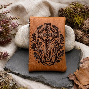 May include: A tan leather zipped pouch featuring a dark brown laser-engraved design of a Celtic cross surrounded by intricate oak leaves and acorns. The rectangular pouch has a visible silver-toned metal zip pull on the left side.