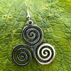 May include: A silver-toned earring featuring a Celtic Triskele design, composed of three interconnected spirals. The earring hangs from a simple hook. The earring is set against a textured green leaf.