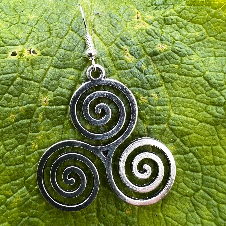 May include: A silver-toned earring featuring a Celtic Triskele design, composed of three interconnected spirals. The earring hangs from a simple hook. The earring is set against a textured green leaf.