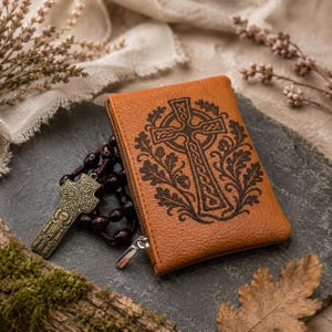 May include: A tan leather zipped pouch featuring a dark brown engraved Celtic cross surrounded by oak leaves and acorns. Beside the pouch lies a rosary with dark brown beads and a metallic bronze-toned crucifix with intricate knotwork patterns.