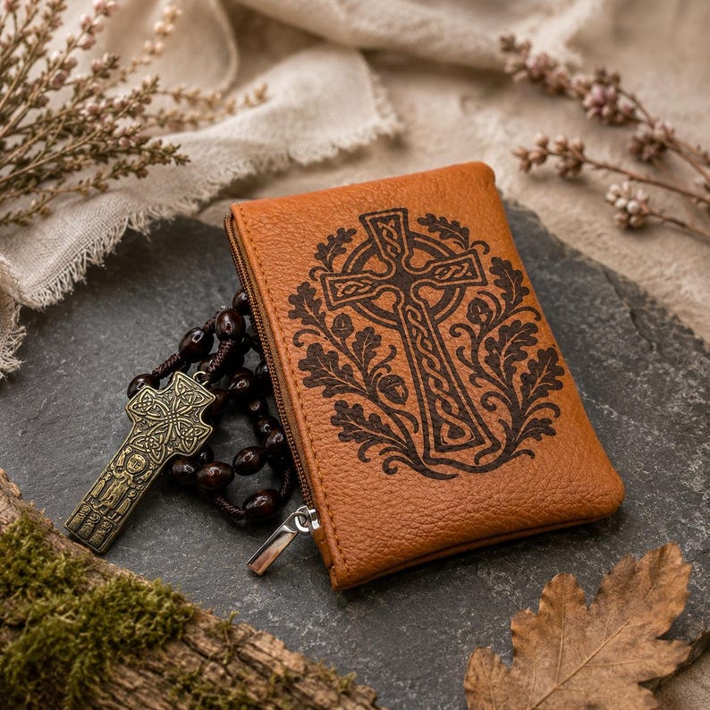 May include: A tan leather zipped pouch featuring a dark brown engraved Celtic cross surrounded by oak leaves and acorns. Beside the pouch lies a rosary with dark brown beads and a metallic bronze-toned crucifix with intricate knotwork patterns.