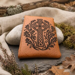 May include: A tan, textured leather pouch featuring a dark, laser-engraved Celtic cross design surrounded by detailed oak leaves and acorns. The rectangular accessory includes a silver-toned metal zip along the left side edge.