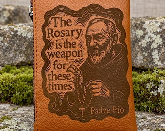 May include: A tan leather zippered pouch featuring a dark brown laser-engraved illustration of Padre Pio holding a rosary. The text reads, The Rosary is the weapon for these times. Padre Pio is written in the lower right corner of the engraving.
