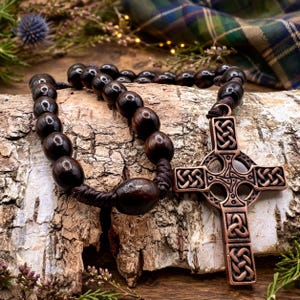 May include: A dark brown wooden rosary with large, rounded beads and a bronze Celtic cross pendant. The rosary is displayed on a textured wooden surface, with a blurred background featuring greenery and a plaid fabric.