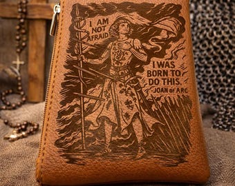 May include: A tan leather pouch with a zip closure, featuring a laser-engraved illustration of Joan of Arc holding a banner. The text reads: I AM NOT AFRAID. I WAS BORN TO DO THIS. JOAN OF ARC. The pouch has a textured surface and a trapezoidal shape.