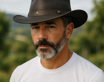 Puede incluir: Un sombrero de vaquero de cuero marrón oscuro con ala ancha y una banda de cuero. El sombrero tiene remaches decorativos y un estilo clásico y robusto. El sombrero lo lleva una persona que lleva una camiseta blanca.